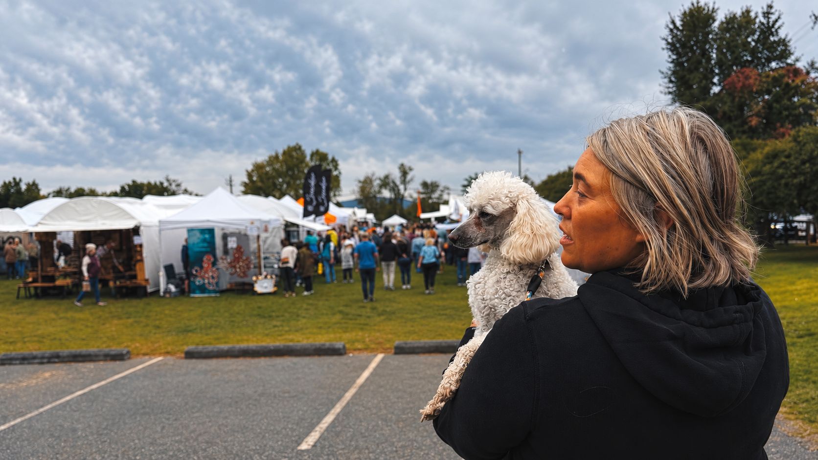 Jessica holding her white poodle at an outdoor craft fair with tents behind her